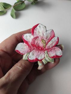 a hand holding a pink and white flower brooch with green leaves in the background