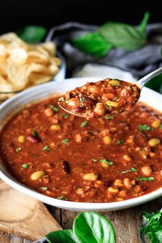 a spoon full of chili and beans with some chips in the background on a wooden table