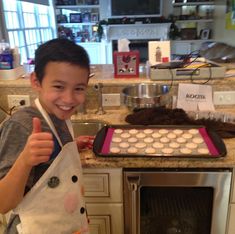 a young boy holding an apron and giving the thumbs up while standing in front of a oven