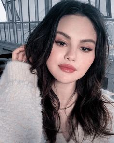 a woman with long dark hair wearing a white sweater and orange lipstick looks at the camera