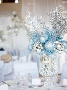 a vase filled with blue and silver ornaments on top of a table covered in white linens