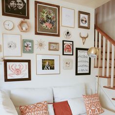 a white couch sitting under a staircase next to a wall with pictures and clocks on it