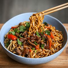 a blue bowl filled with noodles and meat on top of a wooden table next to chopsticks