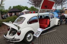 an old white car with its trunk open and people looking at it in the background