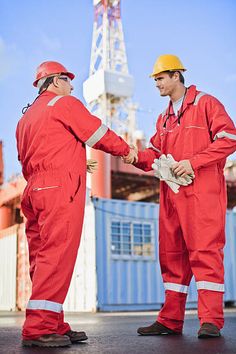 two men in red work suits shaking hands with an oil rig in the back ground