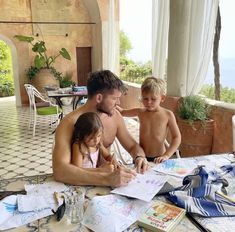 a man and two children sitting at a table in front of an open window with books on it