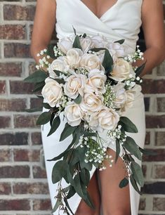 a bridal holding a bouquet of white flowers and greenery in front of a brick wall