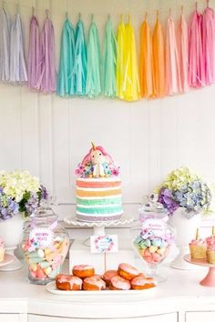 a table topped with lots of cakes and desserts next to rainbow streamers hanging from the ceiling