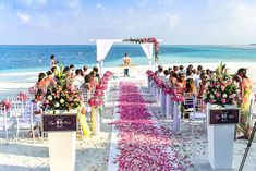 a wedding ceremony on the beach with pink and white flowers lining the aisle, along with people sitting in chairs