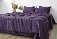 a bed with purple sheets and pillows on top of it in a white walled room