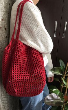 a woman carrying a red crocheted bag in front of a wall with plants