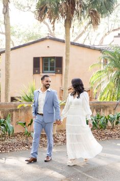 a man and woman holding hands walking in front of a pink house with palm trees