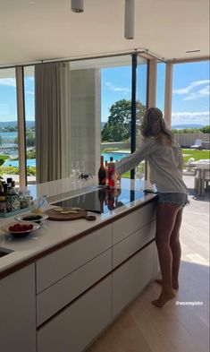 a woman standing at the kitchen counter in front of an open window looking out on water