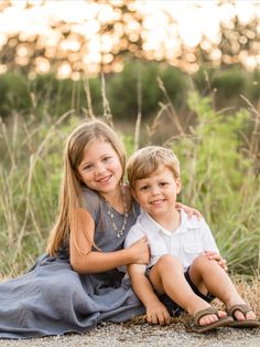 two young children sitting on the ground in front of some tall grass and trees at sunset