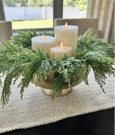 three candles sitting on top of a bowl filled with pine needles and greenery in front of a window