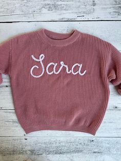 a pink sweater with the word sara written in white across it on a wooden background