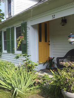 a yellow door sits on the side of a white house with green shutters and plants