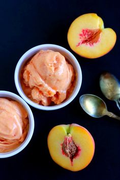 three bowls filled with ice cream next to sliced peaches and spoons on a black surface