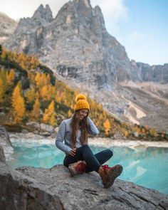 a woman sitting on top of a rock next to a lake wearing a yellow hat