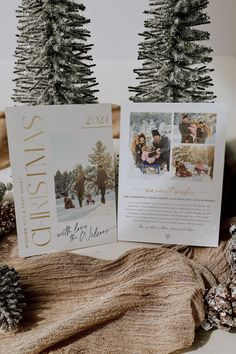 two christmas cards sitting on top of a table next to pine cones and fir trees