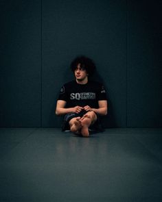 a man sitting on the floor in front of a black wall with his hands crossed