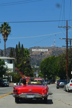 an old red car is driving down the street in front of hollywood sign and palm trees