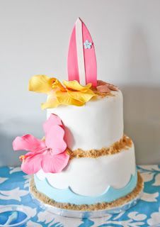 a three tiered cake decorated with flowers and surfboards on the top, sitting on a blue tablecloth