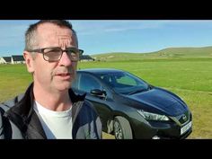 a man wearing glasses standing next to a black car in a green field with hills behind him