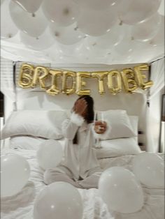 a woman sitting on top of a bed covered in white balloons and gold foil letters