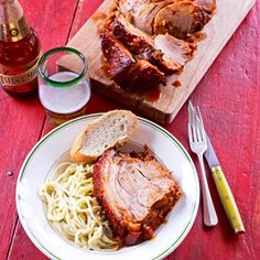 a white plate topped with meat and pasta next to a glass of beer on a red table