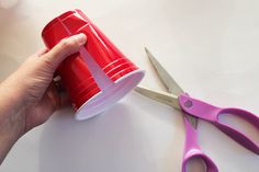 a person holding a red cup with pink handles and scissors on the table next to it