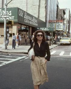 a woman walking across a street in front of a building with people on the sidewalk