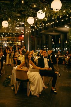 a group of people sitting around each other at a dinner table with lights hanging from the ceiling