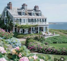 a large house sitting on top of a lush green hillside next to the ocean with lots of flowers in front of it