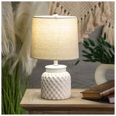 a white table lamp sitting on top of a wooden table next to a book and plant