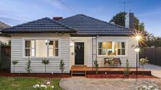 a small white house with a wooden deck in the front yard and landscaping around it