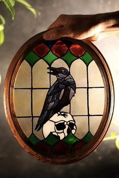 a hand holding a stained glass window with a bird on it's head in front of a tree