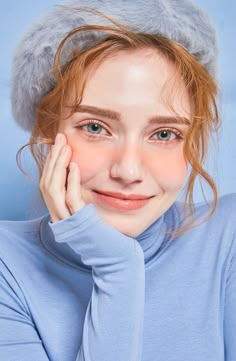 a young woman wearing a blue sweater and a fur hat smiles at the camera with her hand on her cheek