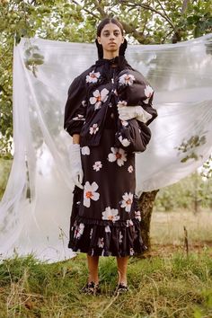 a woman standing in the grass wearing a black dress with flowers on it and white sheer fabric behind her