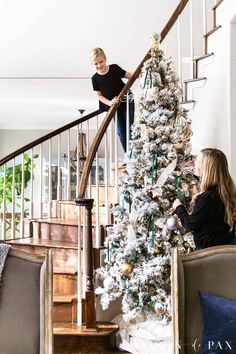 two women sitting at the bottom of stairs near a christmas tree with white and gold decorations