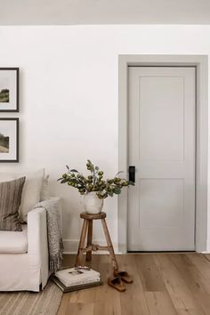 a living room with white walls and wooden flooring, a plant in a vase on a stool next to the door