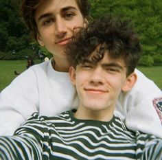 two young men sitting next to each other in a park, one with his head on the back of another man's shoulders