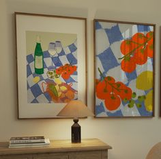 two paintings hang on the wall next to a table with a lamp and bookcase