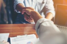 two people shaking hands over a table with papers and pens on it, one person is holding the other's hand