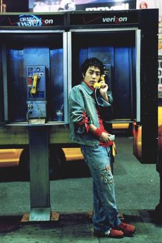 a young man standing in front of a pay phone booth talking on the cell phone