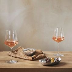 two glasses of wine on a wooden table with some food in the bowl and one glass is empty