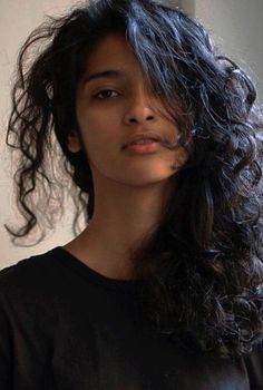 a woman with long dark hair standing in front of a white wall wearing a black t - shirt