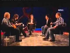 four men sitting in chairs talking to each other on the set of an interview with one man holding a guitar