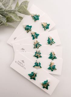 six christmas tree earrings on top of a white card next to a green potted plant