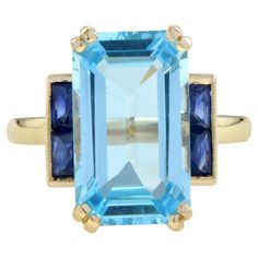 Impressively large and stunning Art Deco style featuring an emerald cut sky blue topaz measuring approx. 15 x 9 mm. or estimate 7.40 carats. Square flat shoulders feature four French cut Sapphires. Truly striking on the finger this ring will not fail to please. Ring Information Style: Art-deco Metal: 14K Yellow Gold Total weight: 4.83 g. (approx. total weight) Center Gemstone Type: Blue Topaz Shape: Emerald Size: 15 x 9 mm. Number: 1 Weight: 7.40 Carat (approx.) Shoulder Gemstones Type: Sapphire French Cut, Sky Blue Topaz, Estilo Art Deco, Art Deco Style, Style Art, Ring Box, Deco Style, Solitaire Ring, Emerald Cut